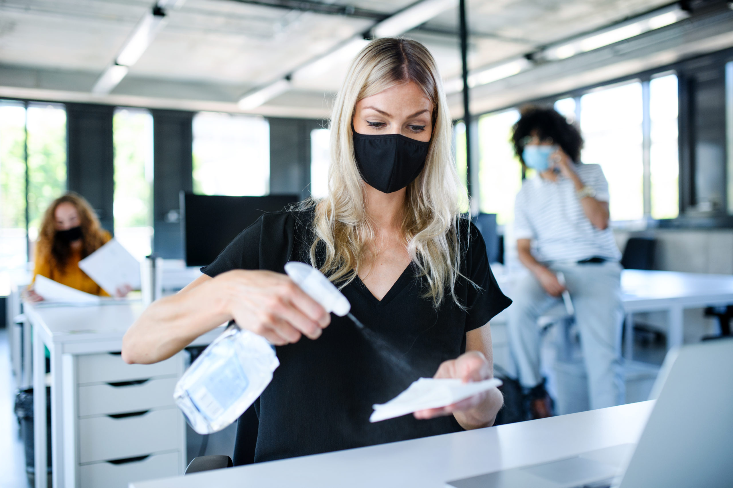 Young woman with face mask back at work in office after lockdown, disinfecting hands.